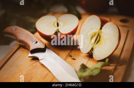 Auf einem hölzernen Schneidebrett liegt ein reifer, köstlicher roter Apfel, in zwei Hälften geschnitten, und daneben liegt ein scharfes Messer. Gesunde Ernährung. Ernährung. Fruit-Har Stockfoto