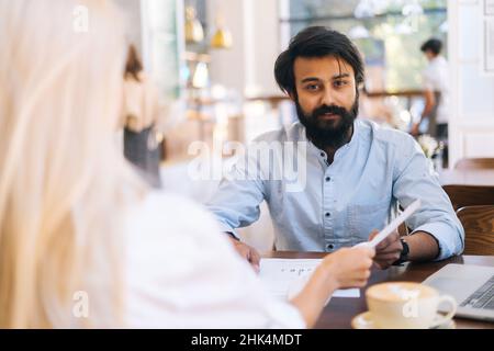 Nahaufnahme einer nicht erkennbaren jungen Geschäftsfrau, die Dokumente an indische Kollegen übergab, im Café am Tisch saß, selektiver Fokus. Stockfoto