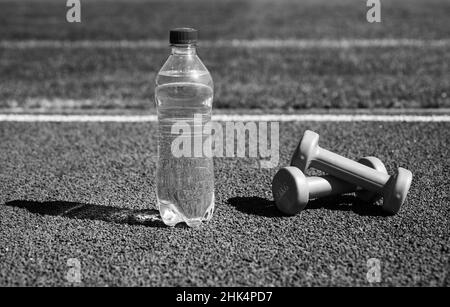 Hydratation nach der Fitness. Gesunder Lebensstil. Wasserhaushalt im Körper beim Training. Langhanteln und Flasche auf der Laufstrecke. Sportgerät und -Ausrüstung Stockfoto