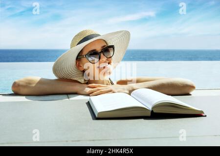 Lächelnde, attraktive Frau mit Hut und Sonnenbrille, die ein Buch liest und sich im unendlichen Swimmingpool im Ferienresort entspannt Stockfoto