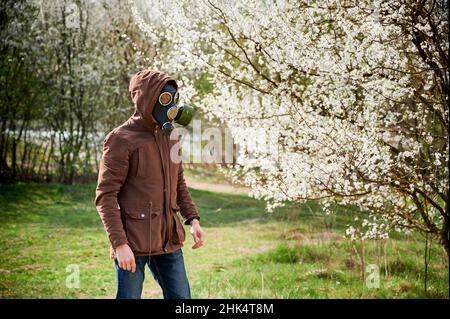 Der Allergiker leidet im Frühjahr an einer saisonalen Allergie, posiert im Frühling im blühenden Garten und trägt eine Gasmaske vor blühenden Bäumen. Frühjahrsallergiekonzept Stockfoto
