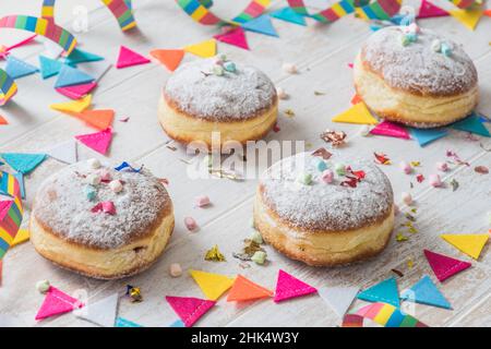 Krapfen, Berliner oder Donuts mit Streamer, Konfetti und Mini-Marschmalchen auf weißem Holzhintergrund. Farbenfrohes Karnevals- oder Geburtstagsbild. Stockfoto