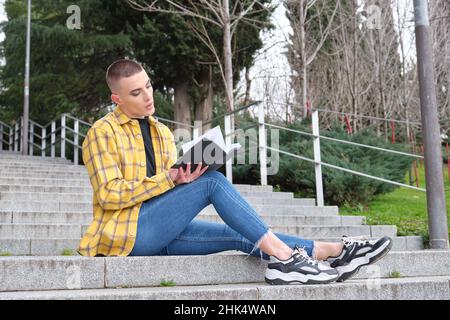 Junge nicht binäre Person, die auf Treppen sitzt und ein Buch liest. Stockfoto