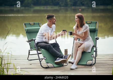 Mann und Frau ruhen sich zusammen auf Stühlen am See aus und trinken Kaffee Stockfoto