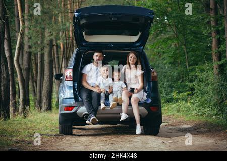 Anreise mit dem Auto Glückliche junge Familienreise zusammen Urlaub. Eltern Vater und Mutter mit niedlichen Kindern in der Nähe sitzen im Kofferraum beim Camping, Picknick Stockfoto