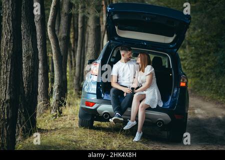 Junges Paar, Mann und Frau, umarmt sich auf einem Picknick, sitzt im Kofferraum eines Autos im Wald, glücklich zusammen Stockfoto