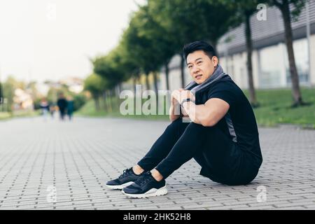 der asiatische Mann nach dem Fitnesstraining und Joggen sitzt müde auf dem Boden und ruht sich aus Stockfoto