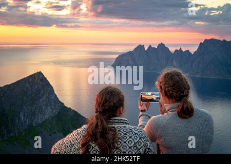 Zwei junge skandinavische Frauen, die den Fjord bei Sonnenuntergang mit dem Smartphone fotografieren, Senja Island, Troms County, Norwegen, Skandinavien, Europa Stockfoto