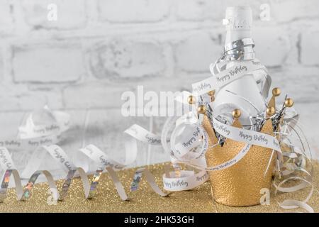 Kleine weiße Flasche mit Sekt in einer dekorativen goldenen Metallkrone, verziert mit Luftschlangen mit Text Happy Birthday, Kopierraum Stockfoto