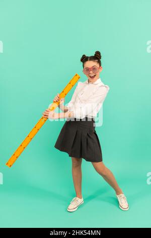 Glückliches Kind in Schuluniform und Brille mit Mathe-Lineal auf blauem Hintergrund, Kindheit Stockfoto