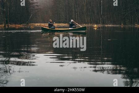 Zwei Männer paddeln mit einem grünen Kanu auf dem Waldsee. Aktiver Lebensstil, wunderschöne Herbst- oder Frühjahrswaldlandschaft Stockfoto