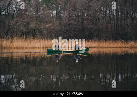 Zwei Männer paddeln ein schönes grünes Kanu auf dem Fluss. Frühlings- oder Herbstszene mit einem Holzboot auf ruhigem Wasser Stockfoto