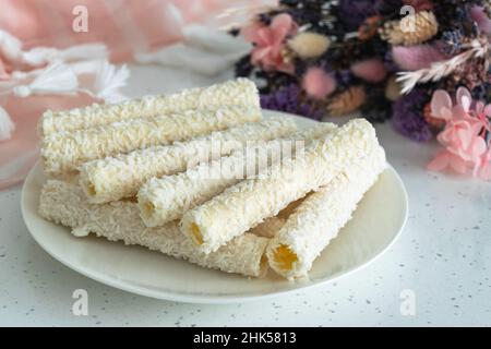 Süßigkeiten für Tee und Kaffee auf einem Porzellanteller. Waffelrollen mit Kokosflocken und Creme neben einem Bouquet von wilden Feldblumen. Horizontaler Phot Stockfoto