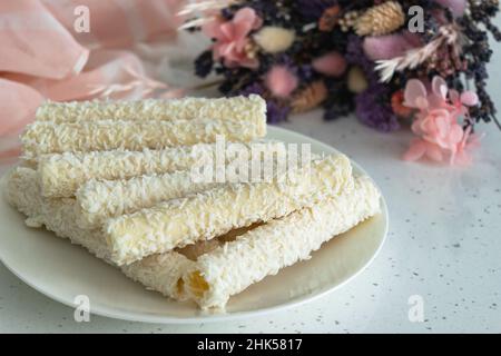Waffelrollen mit Kokosflocken und Creme neben einem Bouquet von getrockneten Blumen. Auf einem hellen Tisch Süßigkeiten für Tee und Kaffee auf einem Porzellanteller. Horizont Stockfoto