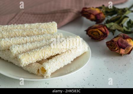 Auf einem hellen Tisch Süßigkeiten für Tee und Kaffee auf einem Porzellanteller. Waffelrollen mit Kokosflocken und Creme neben einem Bouquet aus getrockneten Blumen und einem P Stockfoto