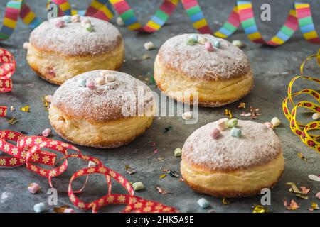 Krapfen, Berliner oder Donuts mit Streamer, Konfetti und Mini-Marschmalchen auf grauem Hintergrund. Farbenfrohes Karnevals- oder Geburtstagsbild Stockfoto