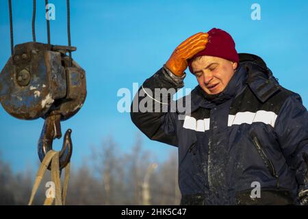 Porträt eines Arbeiters in einer blauen Jacke mit einem Haube am Haken eines LKW-Krans Stockfoto