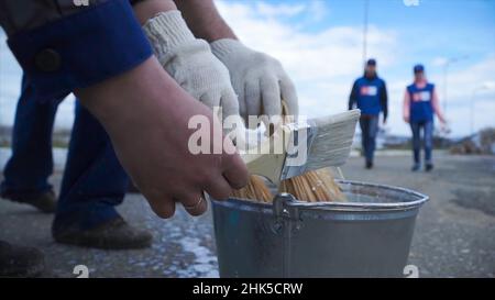 Hände tauchen Farbe in einen vollen Farbbecher. Clip. Nahaufnahme eines Arbeiters seinen Pinsel in die Farbe. Arbeiter in Blau tauchen den Pinsel in die Farbe. Stockfoto