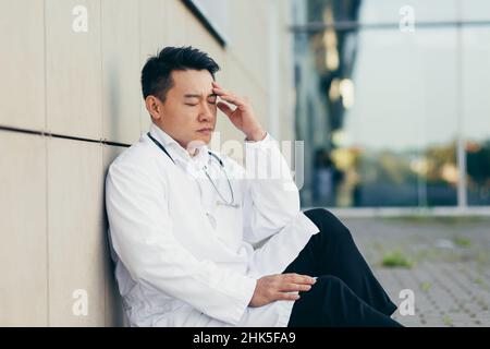 Porträt Arzt Asiatischer Mann müde nach der Arbeit auf dem Boden in der Nähe der Klinik sitzt enttäuscht von dem Ergebnis Stockfoto