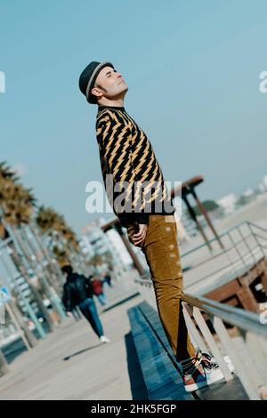 Ein Mann mittleren Alters, der einen braun-schwarz gemusterten Pullover, einen Hut und eine moderne braune Cordhose trägt, steht im Freien, in der Promenade Stockfoto