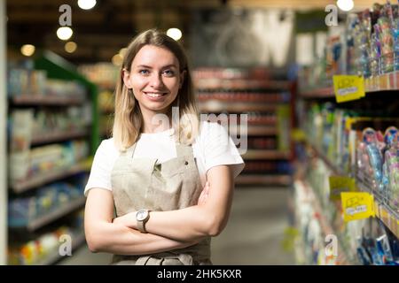 Porträt einer Verkäuferin, einer lächelnden Frau und einem Blick auf die Kamera im Supermarkt. Angenehme freundliche Verkäuferin im Laden zwischen den Reihen stehen. T Stockfoto