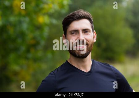 Nahaufnahme Porträt junger, glücklicher bärtiger Mann, der in der Natur zwischen Waldbäumen steht, entspannt, atmet frische Luft und schließt die Augen. Männlich genießt ein Leben von Stockfoto
