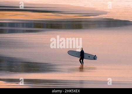 Warmes Abendlicht über einem Surfer, der sein Surfbrett trägt und am Fistral Beach nach einer Surfstunde in Newquay in Cornwall spazierengeht. Stockfoto