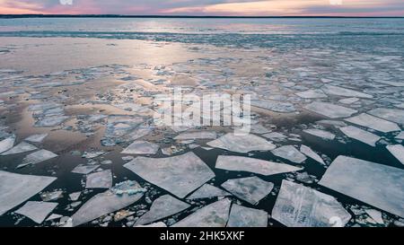 Zerbrochene dicke Eisstücke schwimmen auf einer ruhigen Wasseroberfläche in der Nähe des Flussufers bei hellviolettem Sonnenuntergang am bewölkten Himmel im Winter aus der Luft Stockfoto