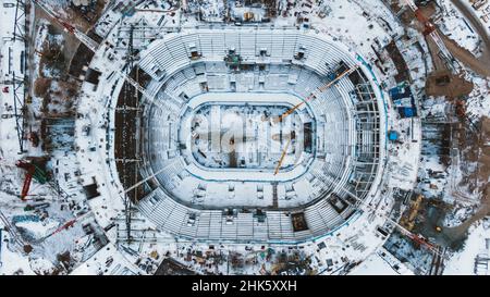 Schneebedeckter Fußballplatz im Stadion mit Plätzen für schneebedeckte Zuschauer und hohen Stahlbaukranen aus der Luft Stockfoto