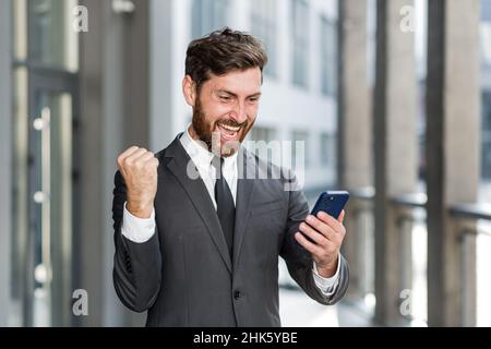 Exited Geschäftsmann feiert Leistung Blick auf Smartphone im Freien. Erfolgreicher Geschäftsmann, der den Sieg mit dem Mobiltelefon im Freien feiert. Frohes Glück Stockfoto