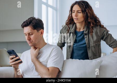 Die junge Frau schaut über die Schulter des Mannes auf den Telefonbildschirm und beginnt zu streiten, den Mann beim Betrug im Wohnzimmer zu beschuldigen Stockfoto