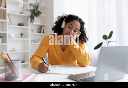 Gelangweilte junge schwarze Studentin, die dumpfe Online-Lektionen auf einem Laptop hat und Informationen zu Hause aufschreibt Stockfoto