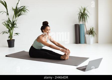 Junge lächelnde Frau beim Stretching-Workout mit Laptop im Yoga-Studio Stockfoto