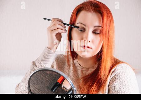 Junge Frau mit roten Haaren trägt Make-up. Lady schaut in den Spiegel und verwendet Kosmetika. Stockfoto