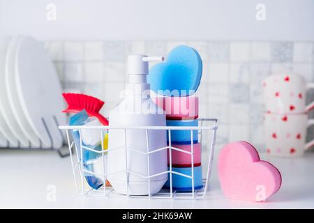 Housekeeping-Konzept Spülmittel, herzförmige Schwämme, Pinsel und Pillen im Korb auf dem Hintergrund von sauberen Tellern und Tassen auf dem Küchentisch. Stockfoto
