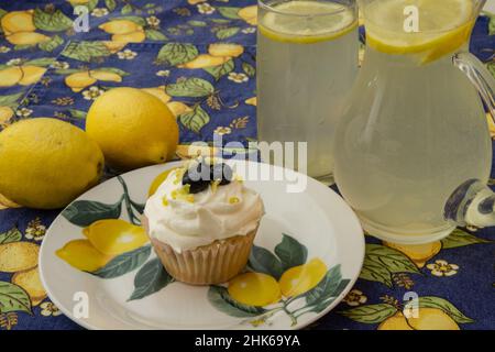 Zitronen-Blaubeer-Cupcakes auf einem Zitronenteller mit Zitronen und Limonade auf einer mit Zitronen gemusterten Tischdecke. Baker - Linda McKusick Stockfoto