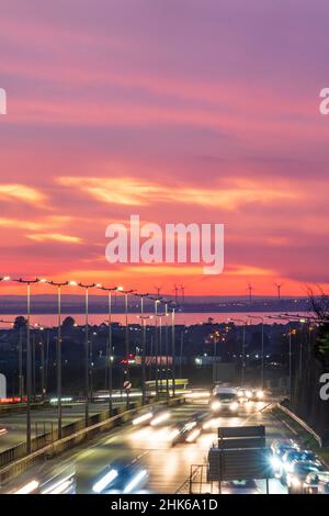 Dramatischer Abendhimmel nach Sonnenuntergang über dem Thanet Way, A299, einer zweispurigen Straße, die North East Kent mit der m2 und London verbindet. Im Hintergrund die Isle of Sheppey, einige Windturbinen und die Themse-Mündung. Rush Hour mit Verkehr auf der Straße. Bewegungsunschärfen mit Scheinwerferspuren. Stockfoto