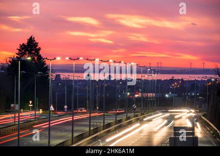 Dramatischer Abendhimmel nach Sonnenuntergang über dem Thanet Way, A299, einer zweispurigen Straße, die North East Kent mit der m2 und London verbindet. Im Hintergrund die Isle of Sheppey, einige Windturbinen und die Themse-Mündung. Rush Hour mit Verkehr auf der Straße. Bewegungsunschärfen mit Scheinwerferspuren. Stockfoto