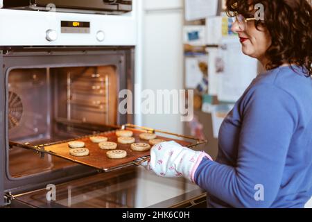 Kaukasische Frau in der Küche, die ein Tablett mit Keksen in den Ofen legte. Backen zu Hause. Selektiver Fokus. Stockfoto