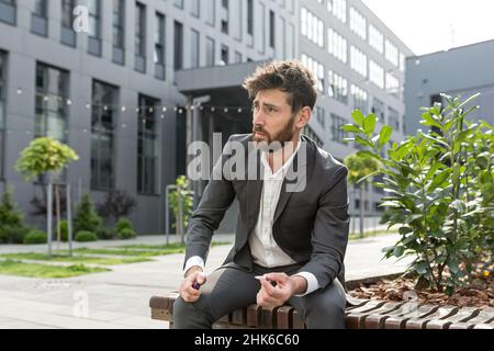 Männlicher Geschäftsmann, der in der Luft in der Nähe des Büros Zigarette raucht, entlastet den Stress nach einem harten Arbeitstag, müde überarbeitet und deprimiert Stockfoto