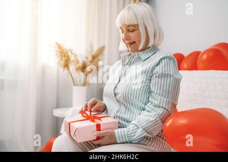 Valentinstag, Muttertag. Ältere blonde Frau 40s 50s Jahre alt trägt stilvolle Bluse offen auf rosa Papier Überraschung Geschenk mit roten Ribbo Stockfoto