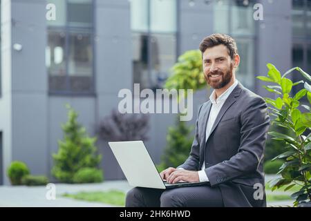 Junger erfolgreicher Geschäftsmann, der am Laptop arbeitet, bärtiger Mann im Business Suit blickt auf die Kamera und lächelt, in der Nähe eines modernen Bürozentrums vor der Tür Stockfoto