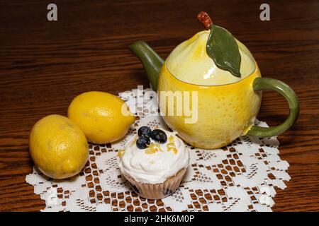 Zitronen-Blaubeer-Cupcakes mit Käsecreme, die auf einem Deckchen mit zwei Zitronen und einer Zitronen-Teekannen zuckend ist. Baker - Linda McKusick Stockfoto