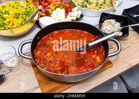 Gulaschsuppe mit Schweinefleisch und verschiedenen Gemüsesorten, in einem Topf mit einem Löffel auf einem mit Lebensmitteln gefüllten Tisch liegend. Stockfoto