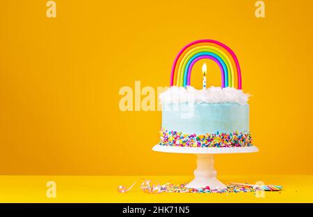 Blauer Geburtstagstorte mit Baiser-Dekorationen, Streuseln und einem farbenfrohen, hausgemachten Regenbogentopfer auf einem fröhlichen gelben Hintergrund. Stockfoto