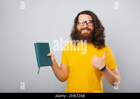 Der kluge junge Mann zeigt den Daumen hoch, während er sein Tagebuch hält. Hochformat auf grauem Hintergrund. Stockfoto