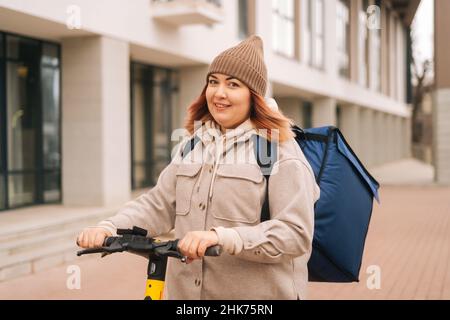 Freundliche Kurierin mit großem Thermorucksack, der mit einem Elektroroller in der Stadtstraße steht und die Kamera anschaut. Stockfoto