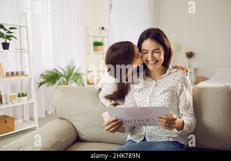 Die kleine Tochter küsst ihre Mutter, während sie ihre DIY-Grußkarte am Muttertag gibt Stockfoto