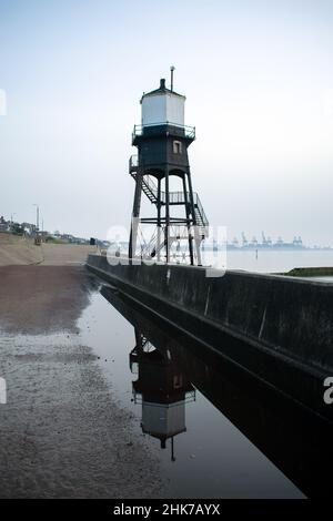 Der Dovercourt Lighthouse spiegelt sich an einem bewölkten und nebligen Tag in einer Pfütze auf der Promenade wider. Im Hintergrund ist der Hafen von Felixstowe zu sehen. Stockfoto