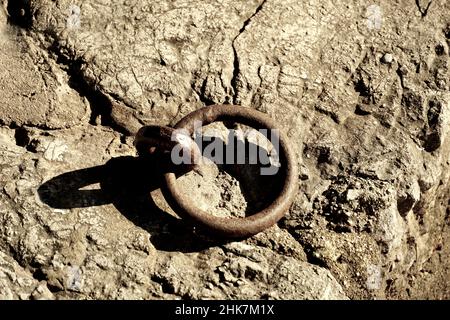 Rostiger, alter, kupferfarbener Rundhaken aus Eisen zum Festmachen von Schiffen und Booten auf dem Hintergrund eines von der Sonne erhellten rauen Steins Stockfoto